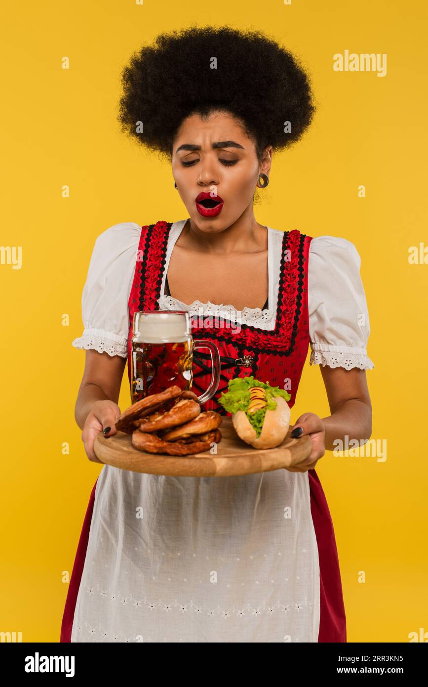 Staunte afroamerikanische bayerische Kellnerin mit Bier und köstlichen Leckereien auf Holztablett auf gelb Stockfoto