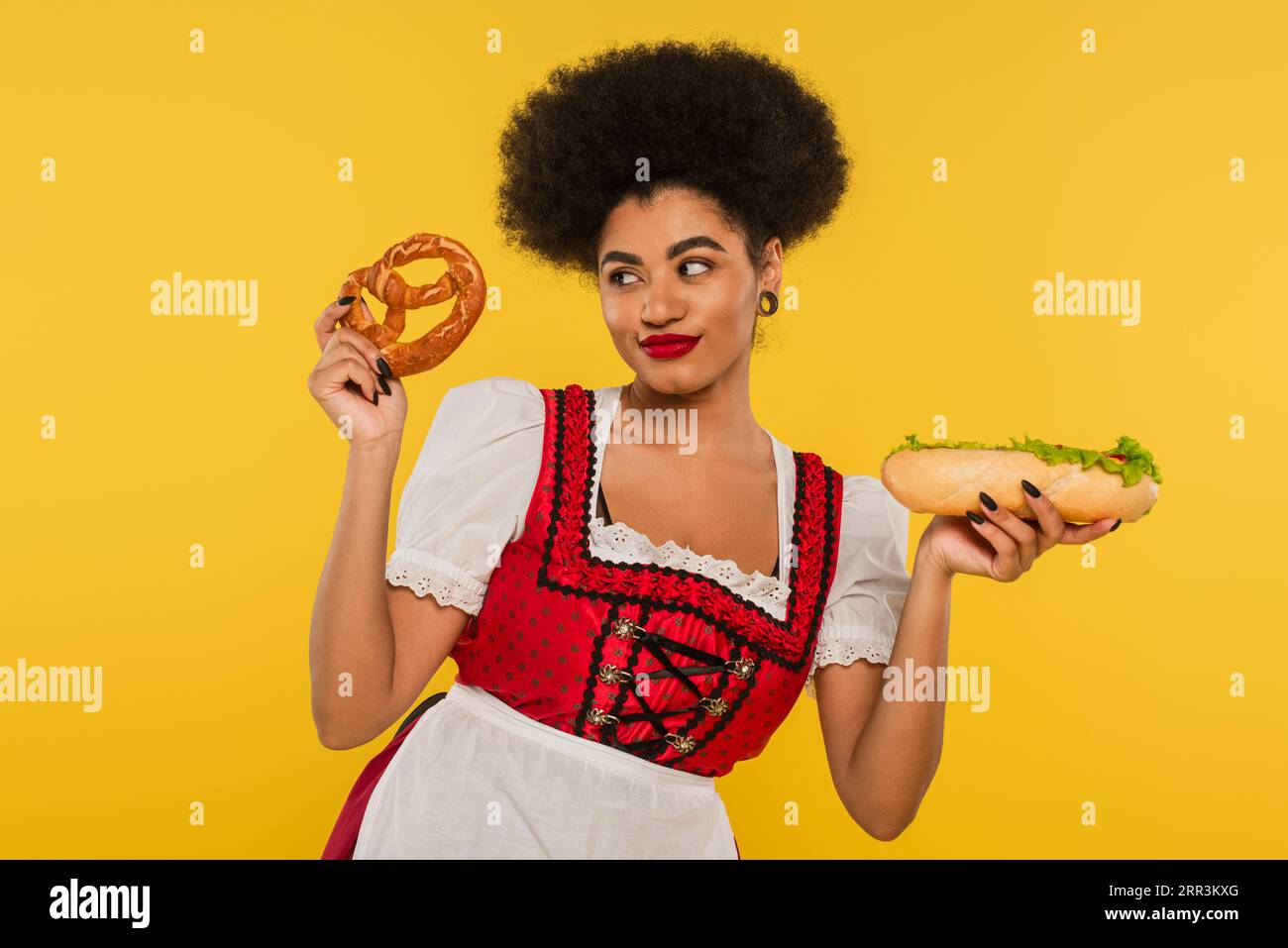 Fröhliche afroamerikanische oktoberfest-Kellnerin in traditionellem Kleid mit Hot Dog und Brezel auf gelb Stockfoto