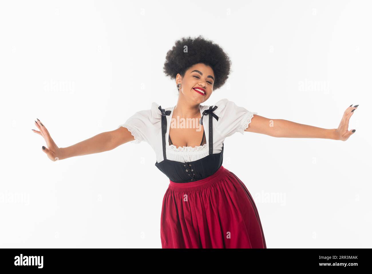 Fröhliche afroamerikanische Kellnerin in traditioneller bayerischer Tracht mit ausgestreckten Händen auf weiß Stockfoto