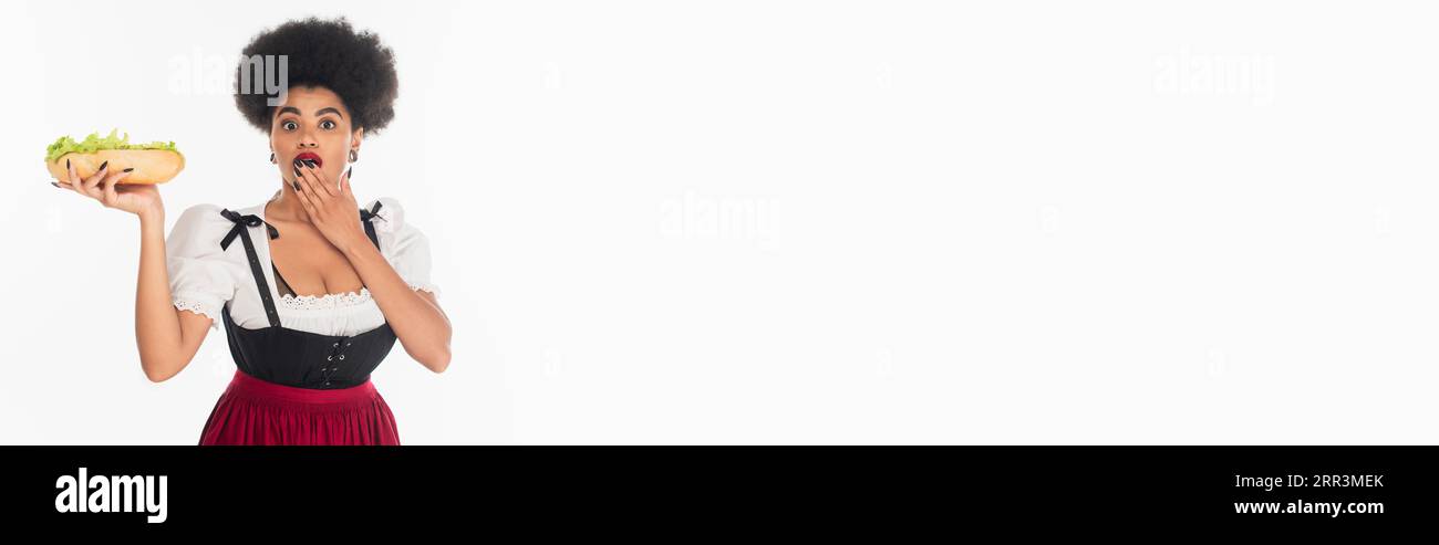 Staunende afroamerikanische oktoberfest-Kellnerin mit Hot Dog, der den Mund mit der Hand auf weißem Banner bedeckt Stockfoto