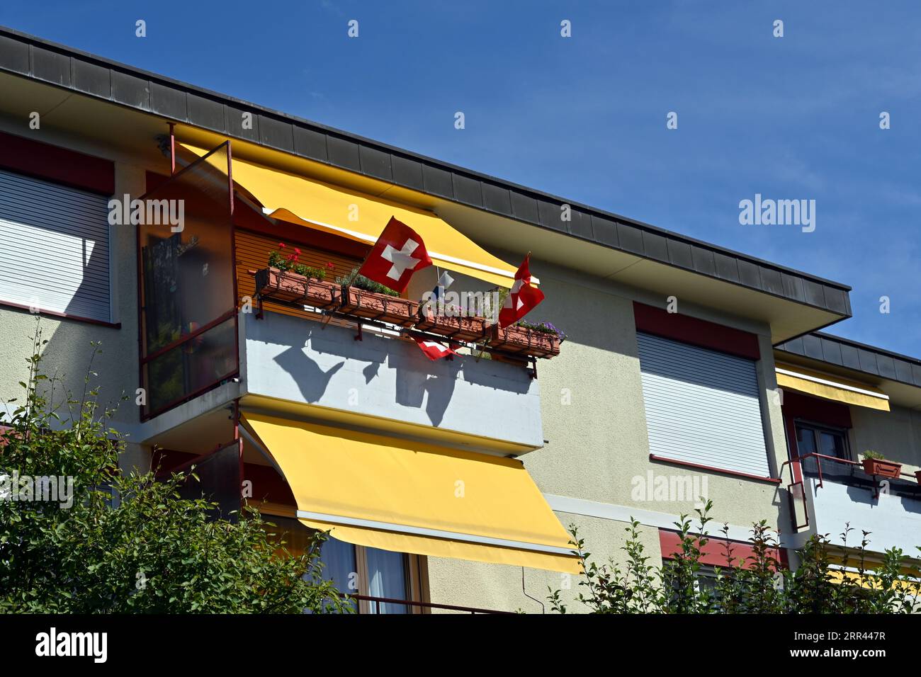 Schweizer Fahnen wehen im Wind. Sie sind auf einem Balkon eines alten traditionellen Schweizer Hauses unter gelben Sonnenjalousien angebracht. Stockfoto