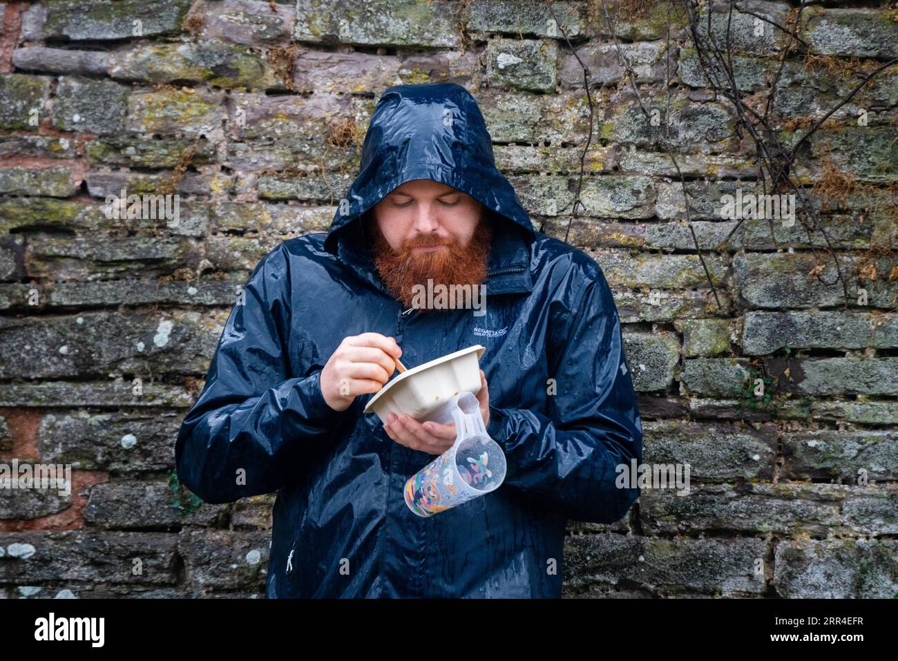 Ein nasser, bärtiger Mann in nassen Regenkleidung mit einem Bierglas isst Fast Food. Green man Festival, Brecon, Wales, Großbritannien, 2023. Foto: Rob Watkins Stockfoto