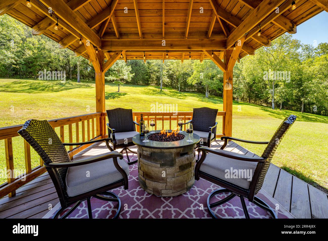 Eine Wohnszene im Freien mit vier Stühlen, die einen Feuerstein unter einem Pavillon in einem Landhaus umgeben. Eine Weinflasche und stielförmige Gläser sitzen am Feuer. Stockfoto