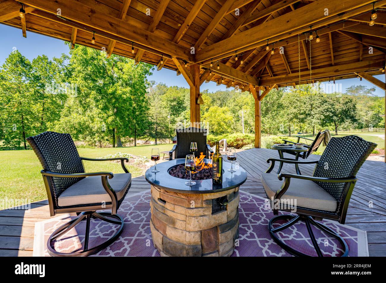 Eine Wohnszene im Freien mit vier Stühlen, die einen Feuerstein unter einem Pavillon in einem Landhaus umgeben. Eine Weinflasche und stielförmige Gläser sitzen am Feuer. Stockfoto
