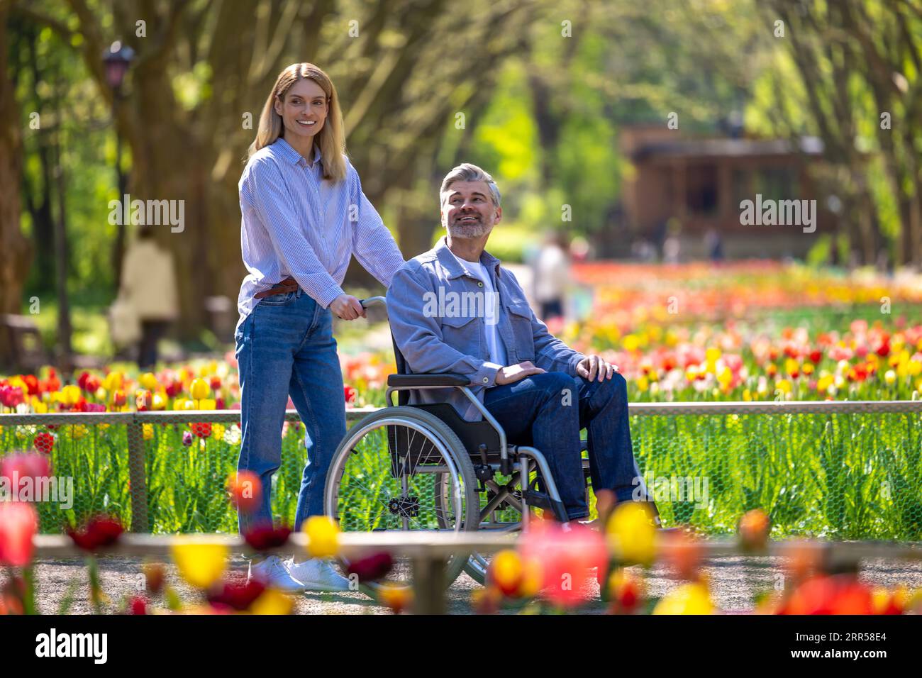 Ehefrau mittleren Alters und behinderter Ehemann im Rollstuhl, der im Park spaziert. Stockfoto