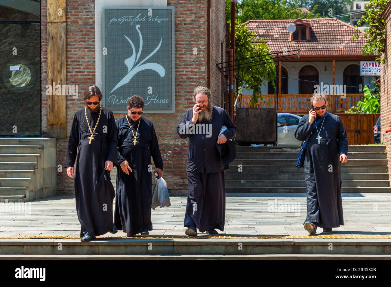 Vier orthodoxe Geistliche in schwarzen Roben gehen über einen gepflasterten Platz in Mtskheta, Region Mtskheta-Mtianeti, Georgien, nahe der Svetitskhoveli-Kathedrale (georgisch სვეტიცხოვლის საკათედრო ტაძარი, wörtliche Übersetzung des georgischen Namens: Kathedrale des Lebensspendenden Pfeilers). Drei von ihnen tragen ein Brustkreuz an einer Kette. Zwei von ihnen telefonieren mit ihren Handys. Einer trägt eine Plastiktüte. Hinter ihnen hängt an einer Ziegelmauer ein Schild mit georgischer Kursivschrift und den englischen Worten „Wedding House“. Im Hintergrund steht ein Haus mit einem hölzernen Balkon. Die Kathedrale wurde zwischen 1010 und 1029 erbaut. Seit 1 Stockfoto
