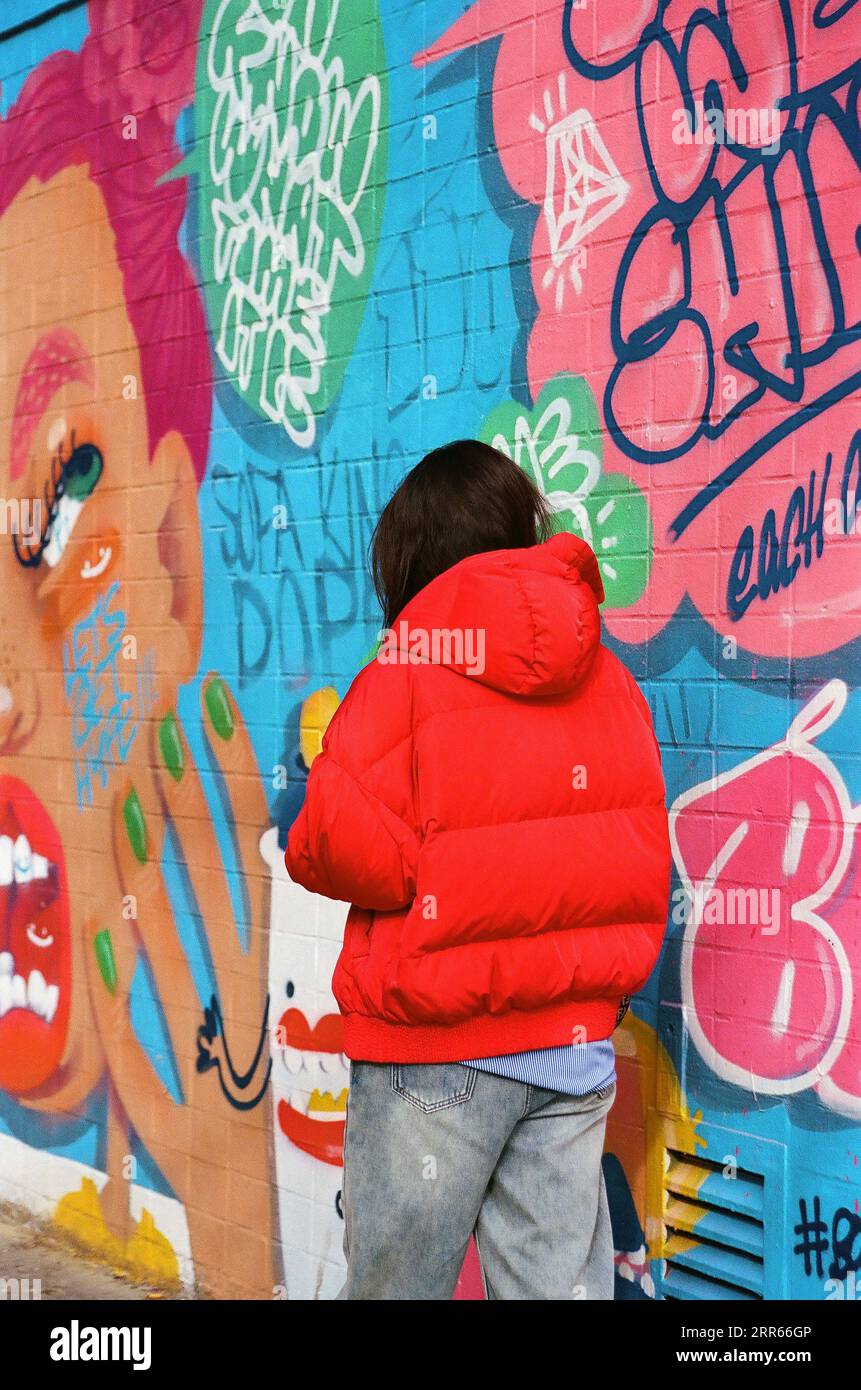 Rückansicht einer Person in rotem Pufferkleid, die an einer Wand mit Graffiti und Kunstwerken vorbeigeht Stockfoto