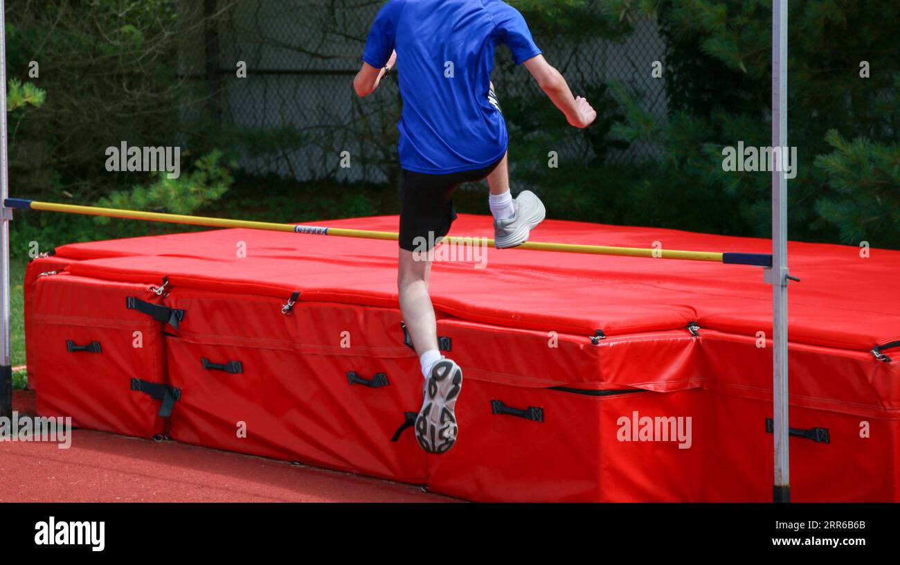 Rückansicht eines jungen männlichen Athleten, der über eine gelbe Hochsprungstange auf rote Matten springt. Stockfoto