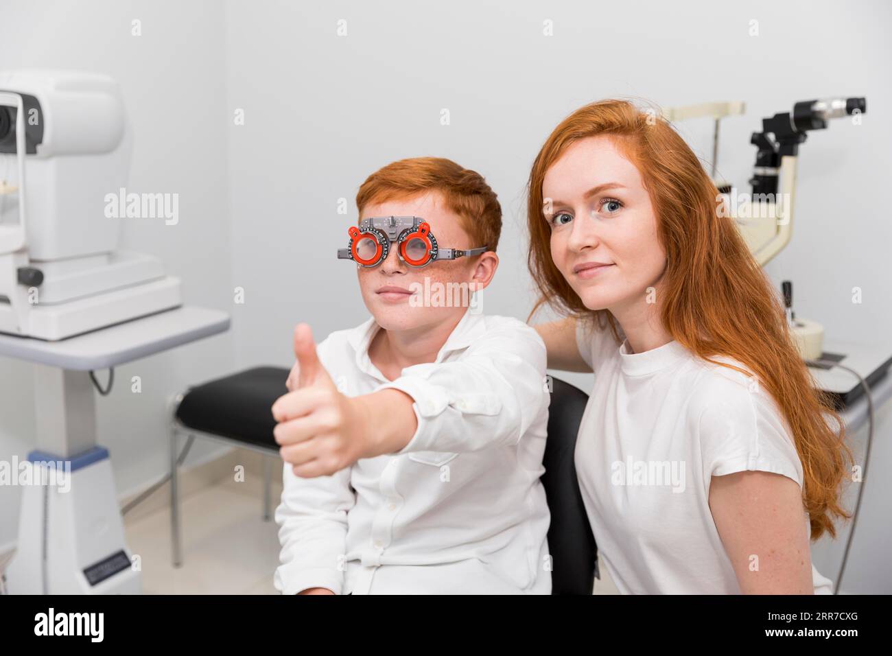 Junge mit Optometrist-Versuchsrahmen mit Daumen-hoch-Geste sitzend mit junger Augenärztin Stockfoto