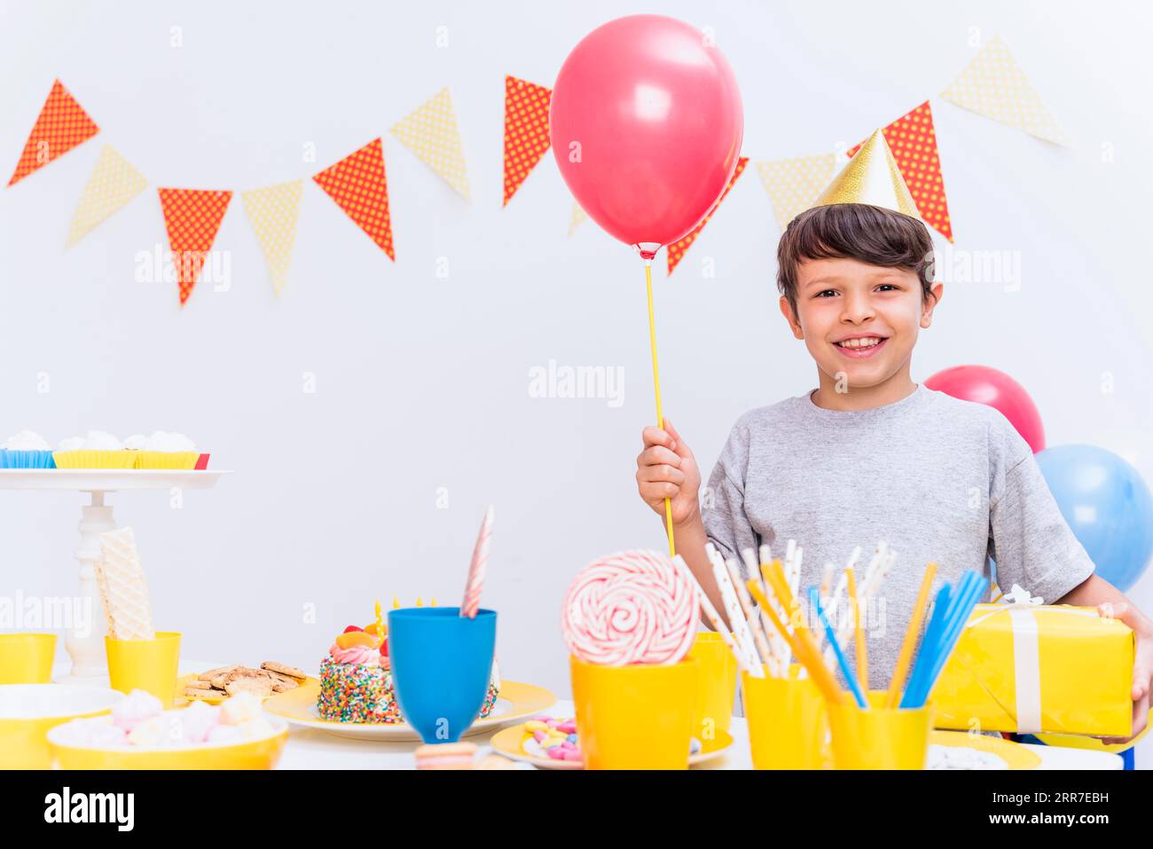 Lächelnder Junge, der einen Party-Hut trägt, der Ballongeschenk hält, der Varieté-Esstisch steht Stockfoto