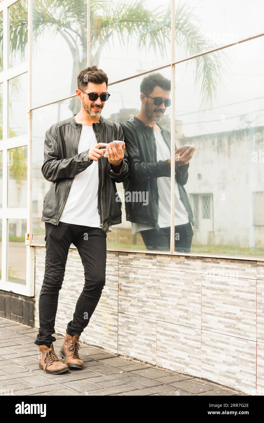 Hübscher junger Mann, der mit dem Smartphone an die Glaswand lehnt Stockfoto