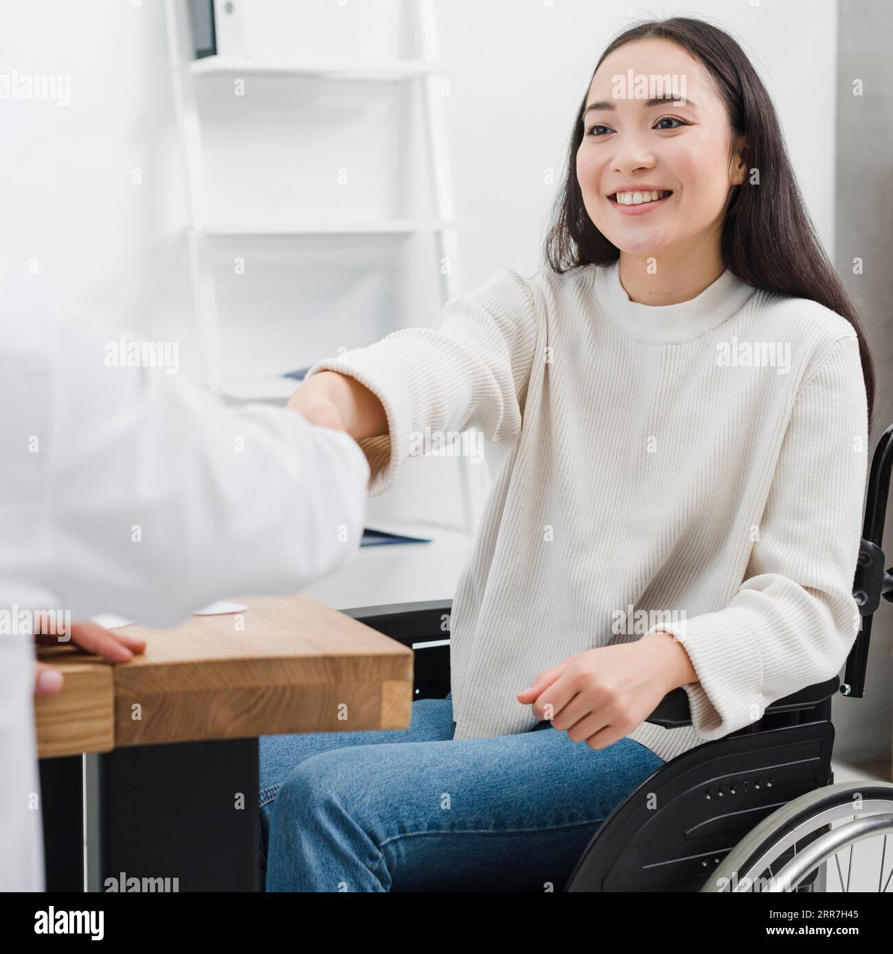 Lächelndes Porträt behinderte junge Frau, die im Rollstuhl sitzt und mit der Person am Arbeitsplatz die Hände schüttelt Stockfoto