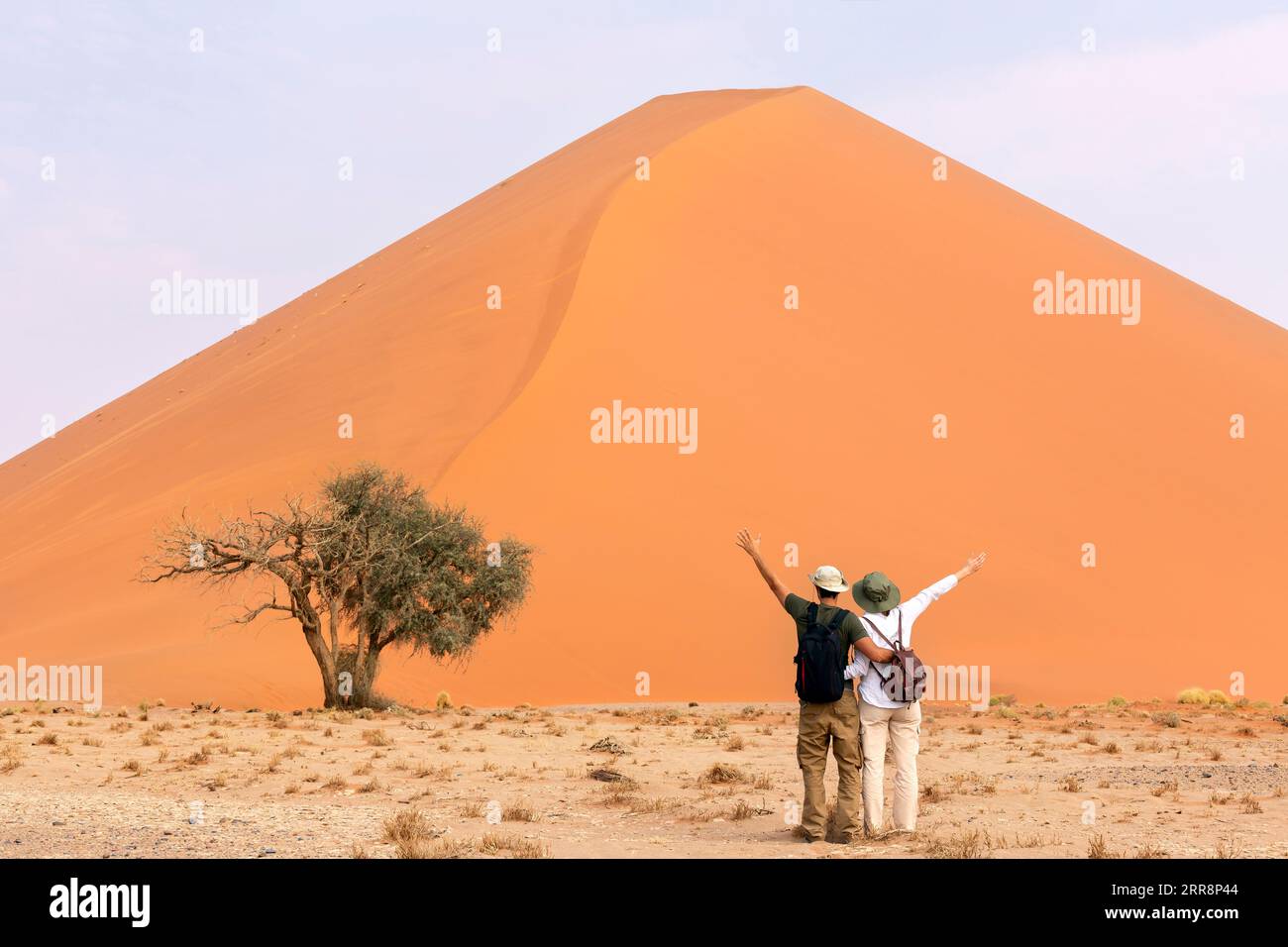 Ein Paar Reisende, die in der Nähe der orangefarbenen Sanddüne mit offenen Armen stehen. Reisewüstenkonzept Stockfoto