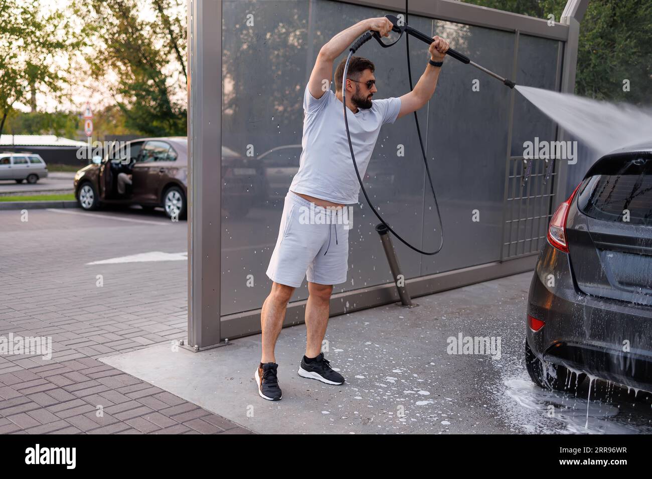 Bei einer Autowaschanlage wäscht ein Mann den Schaum ab, indem er ihn mit einer Wasserkanone über den Kopf hebt. Ein Auto in einer Selbstbedienungswaschanlage. Stockfoto