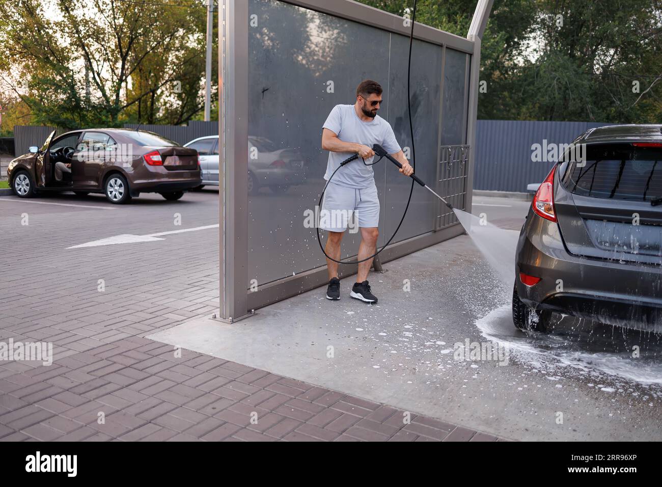 Bei der Autowaschanlage wäscht ein Mann den Schaumstoffschaum mit einer Wasserkanone von der Unterseite des Fahrzeugs. Ein Auto in einer Selbstbedienungswaschanlage. Stockfoto