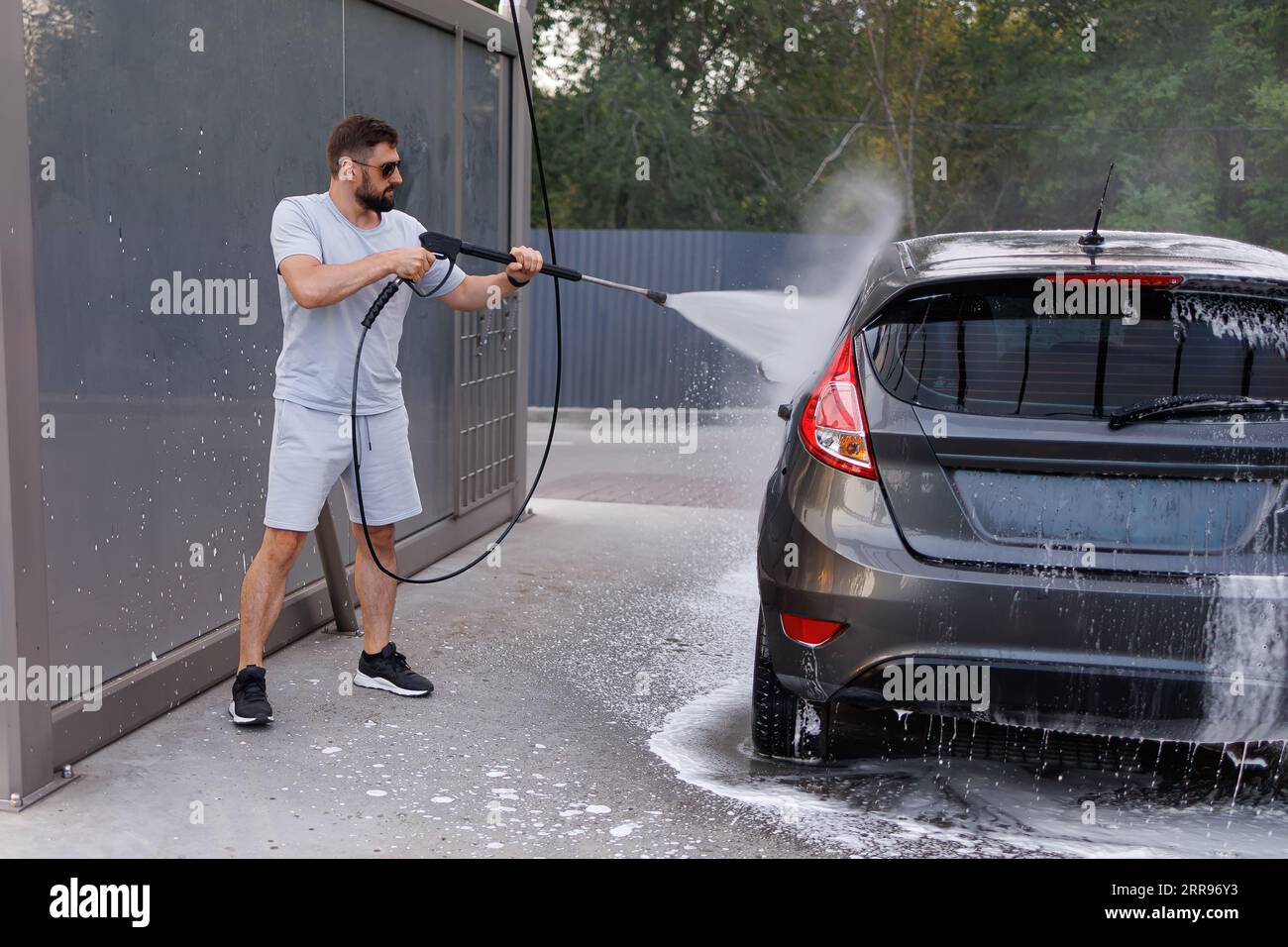 Der Mann hat eine Wasserkanone in der Hand, mit der sie das Auto wäscht. Ein Auto in einer Selbstbedienungswaschanlage. Stockfoto