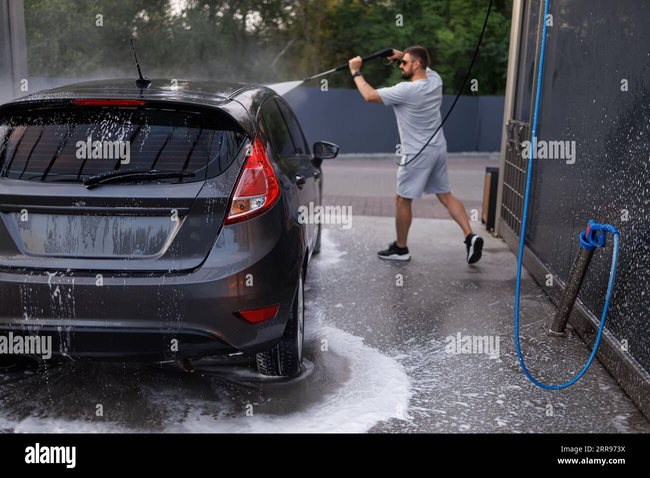 Das Auto an der Autowaschanlage, die im Hintergrund von einem Mann mit einer Wasserkanone gewaschen wird. Ein Auto in einer Selbstbedienungswaschanlage. Stockfoto
