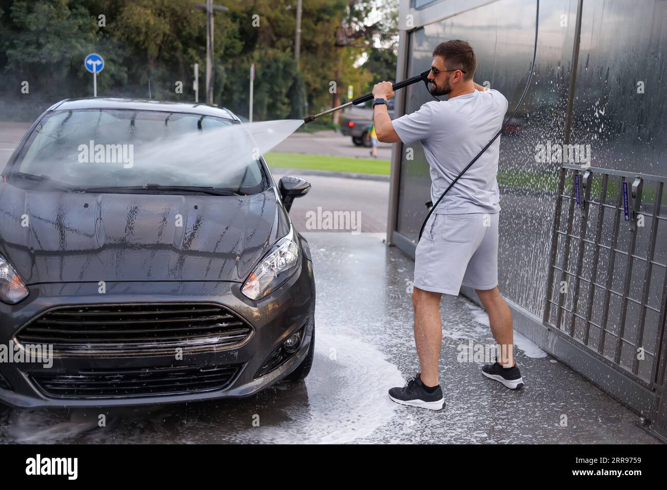 Ein Mann zeigt eine Wasserkanone auf die Windschutzscheibe eines Autos. Ein Auto in einer Selbstbedienungswaschanlage. Stockfoto