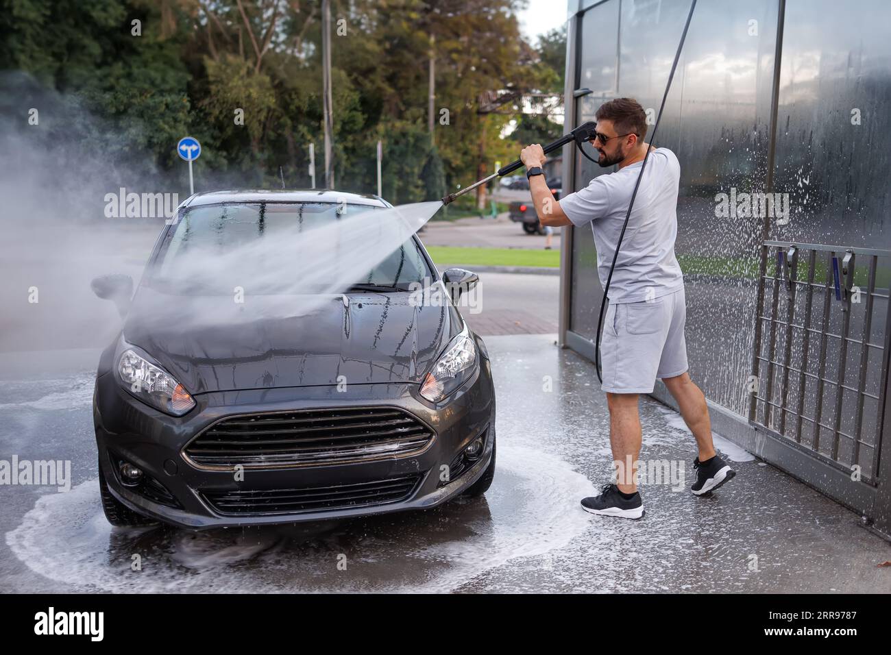 Ein Mann zeigt eine Wasserkanone auf die Motorhaube eines Autos. Ein Auto in einer Selbstbedienungswaschanlage. Stockfoto