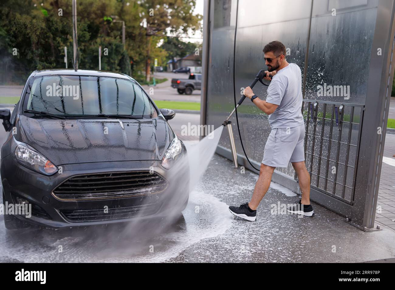 Der Mann richtet die Wasserkanone auf die Frontscheinwerfer des Fahrzeugs und wäscht sie. Ein Auto in einer Selbstbedienungswaschanlage. Stockfoto