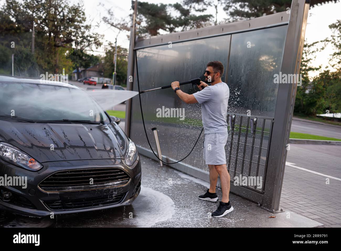 Ein Mann an der Waschanlage zeigt eine Wasserkanone auf die Windschutzscheibe. Ein Auto in einer Selbstbedienungswaschanlage. Stockfoto
