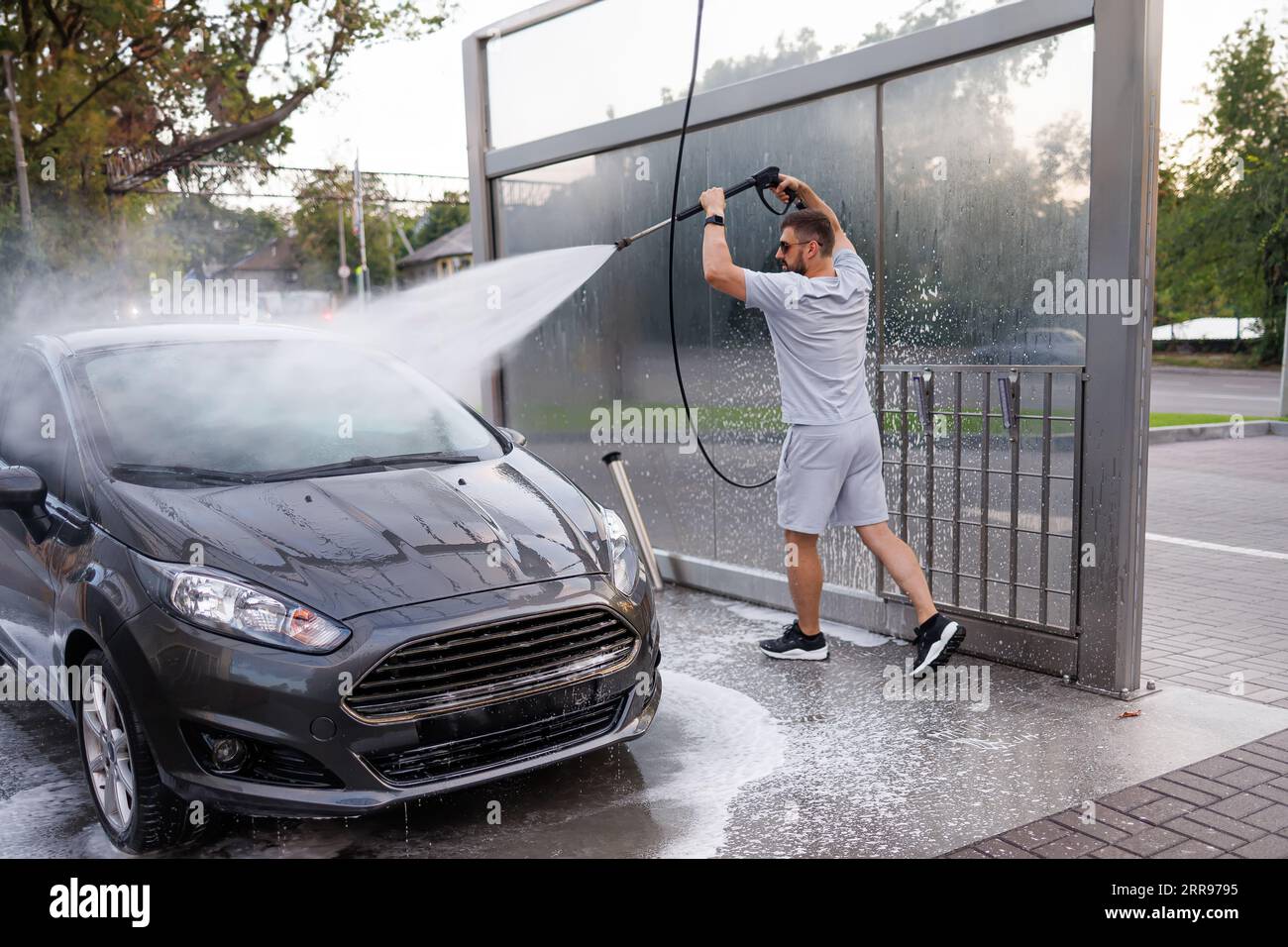 Ein Mann an der Autowaschanlage peitscht die Wasserkanone über seinen Kopf und richtet sie auf die Windschutzscheibe. Ein Auto in einer Selbstbedienungswaschanlage. Stockfoto