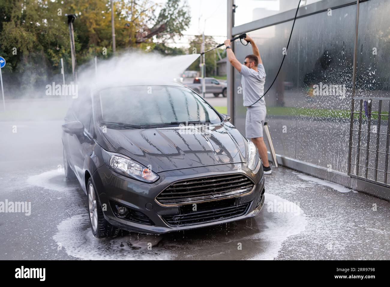 Der Mann im Hintergrund hat die Wasserkanone über seinen Kopf gehoben und zielt auf das Dach des Autos. Ein Auto in einer Selbstbedienungswaschanlage. Stockfoto