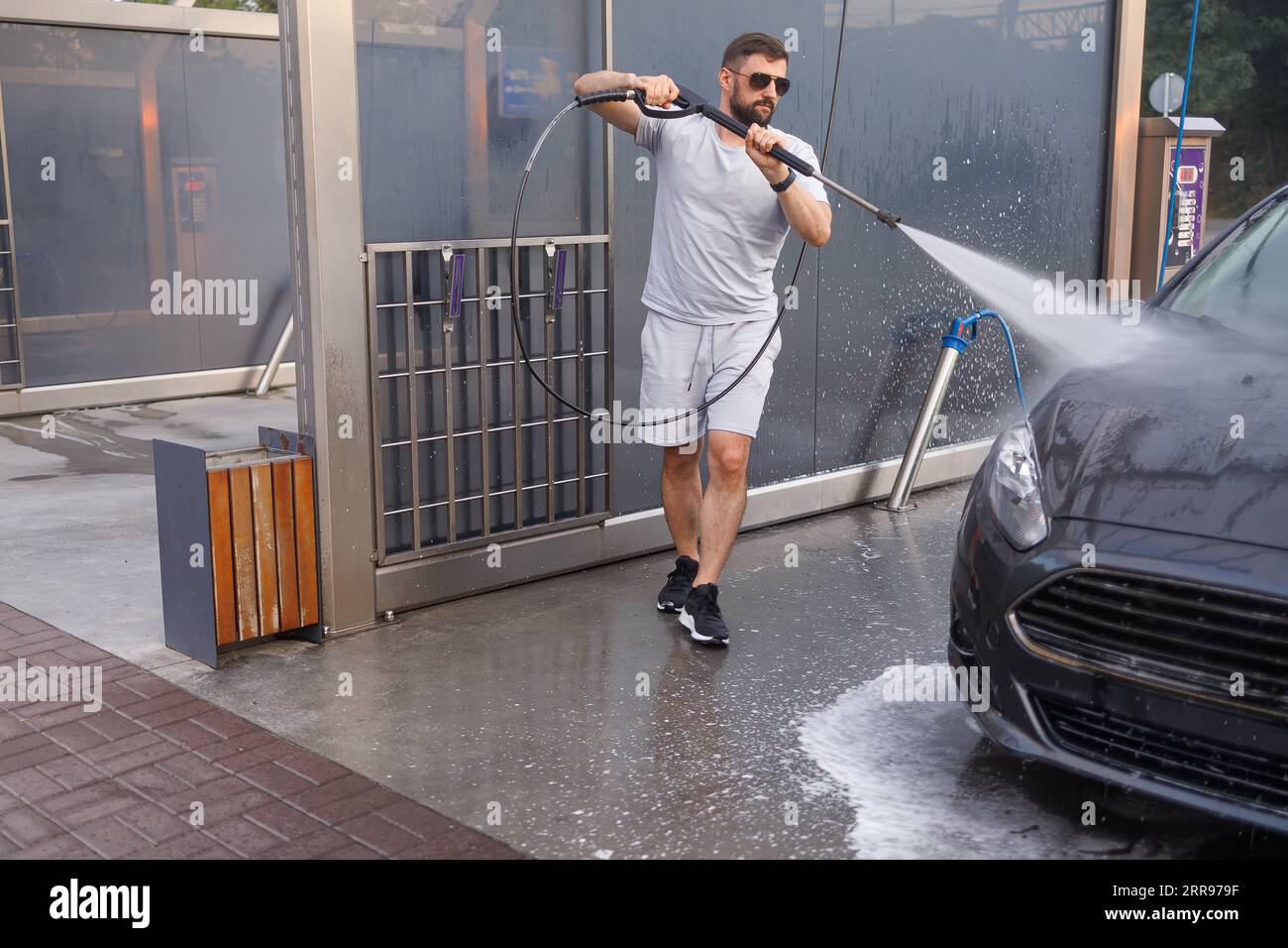 Ein Mann mit einer Wasserkanone in der Hand läuft um das Auto herum und wäscht es. Ein Auto in einer Selbstbedienungswaschanlage. Stockfoto