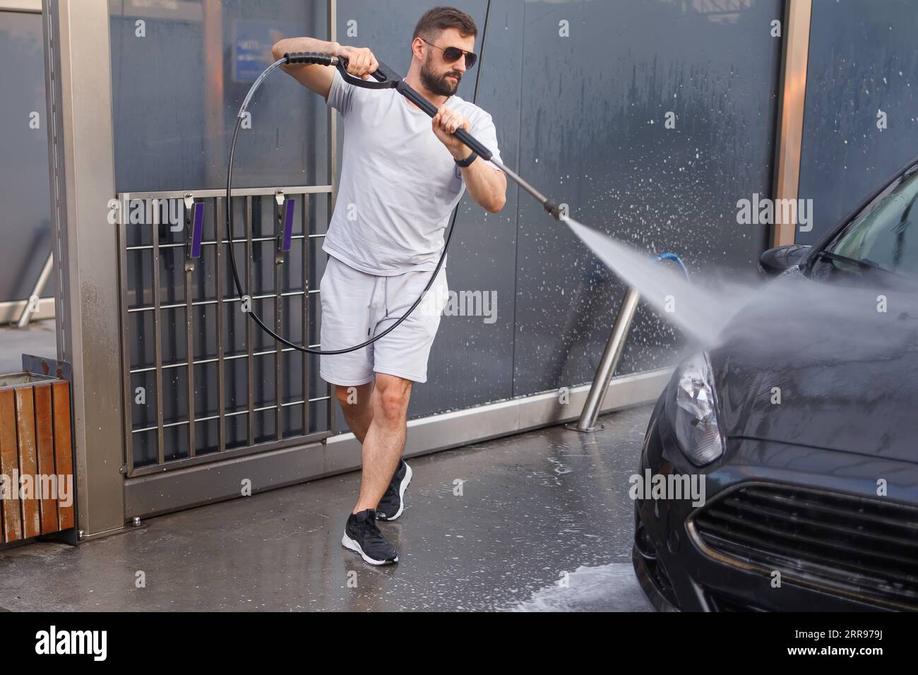 Ein Mann mit einer Wasserkanone in der Hand läuft um das Auto herum und wäscht es. Ein Auto in einer Selbstbedienungswaschanlage. Stockfoto