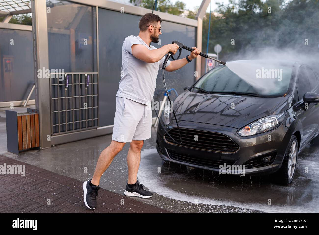 Ein Mann steht vor dem Auto und zeigt mit einer Wasserkanone darauf und wäscht das Wasser ab. Ein Auto in einer Selbstbedienungswaschanlage. Stockfoto