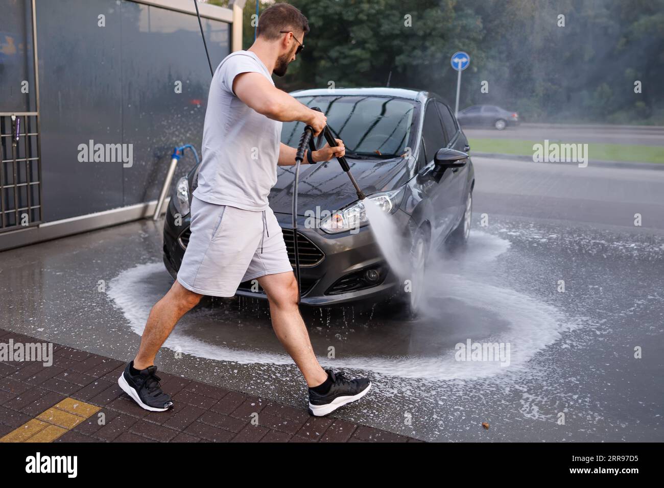 Ein Mann steht vor dem Auto und zeigt mit einer Wasserkanone darauf und wäscht das Wasser ab. Ein Auto in einer Selbstbedienungswaschanlage. Stockfoto