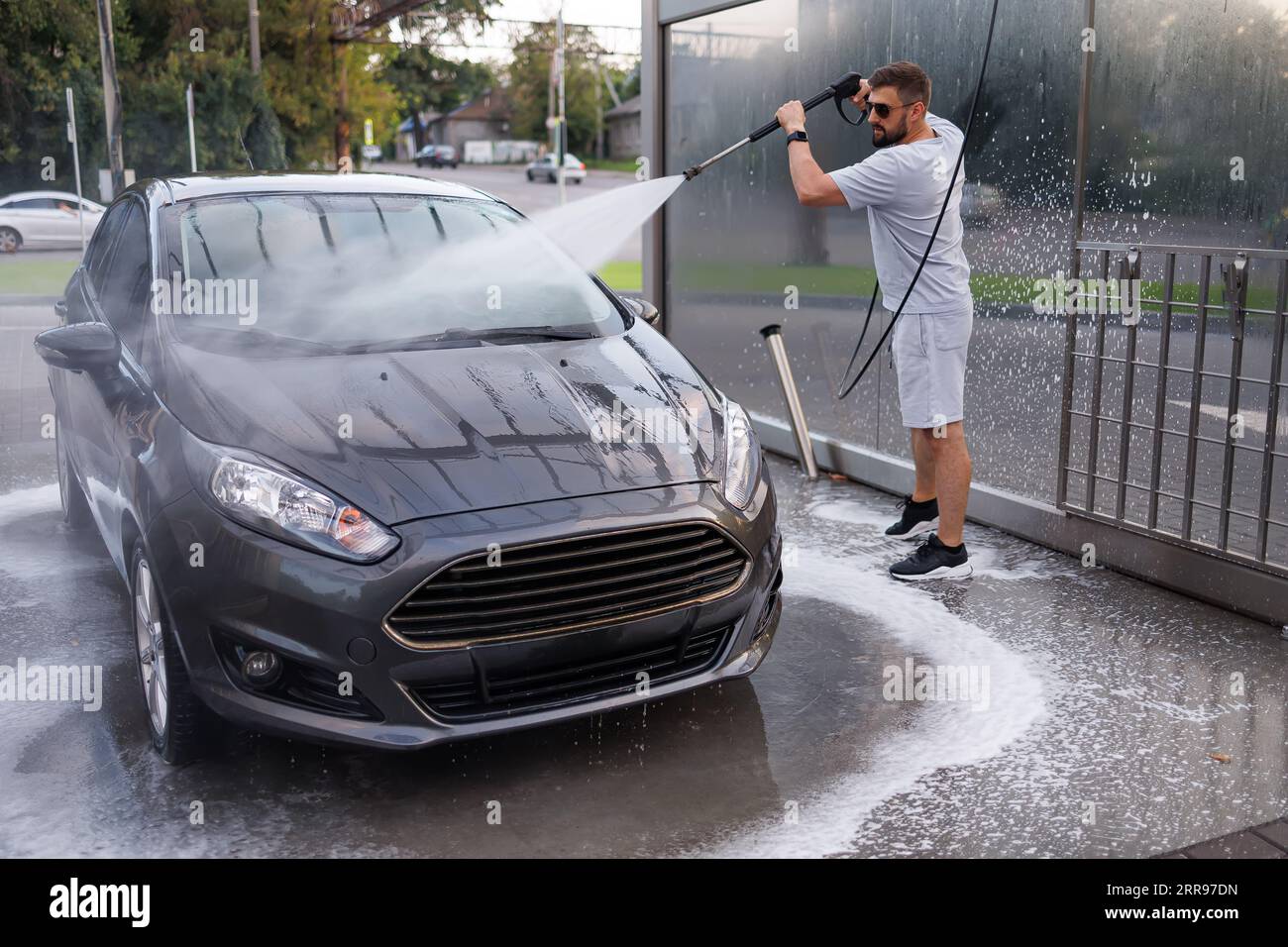 Ein Mann reinigt den Schmutz gründlich mit einer Wasserkanone vom Auto. Ein Auto in einer Selbstbedienungswaschanlage. Stockfoto