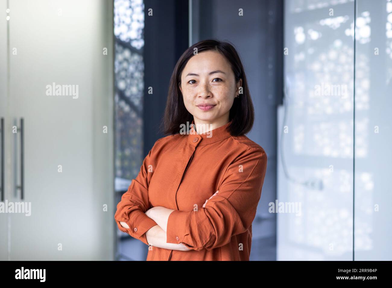 Porträt einer jungen, schönen, selbstbewussten asiatischen Geschäftsfrau, die in einem Bürozentrum steht, mit gekreuzten Armen auf der Brust und ernsthaft auf die Kamera schaut. Stockfoto