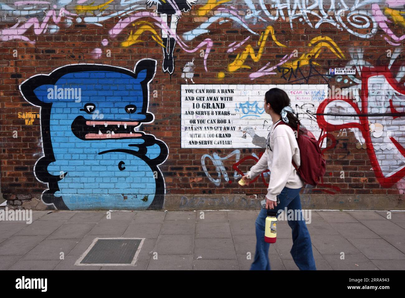 Junge Frau, die an Grafitti oder Wandmalerei vorbeigeht und mit Versen oder Slogan über die Arbeit von Banksy im Ostseedreieck Liverpool spricht Stockfoto