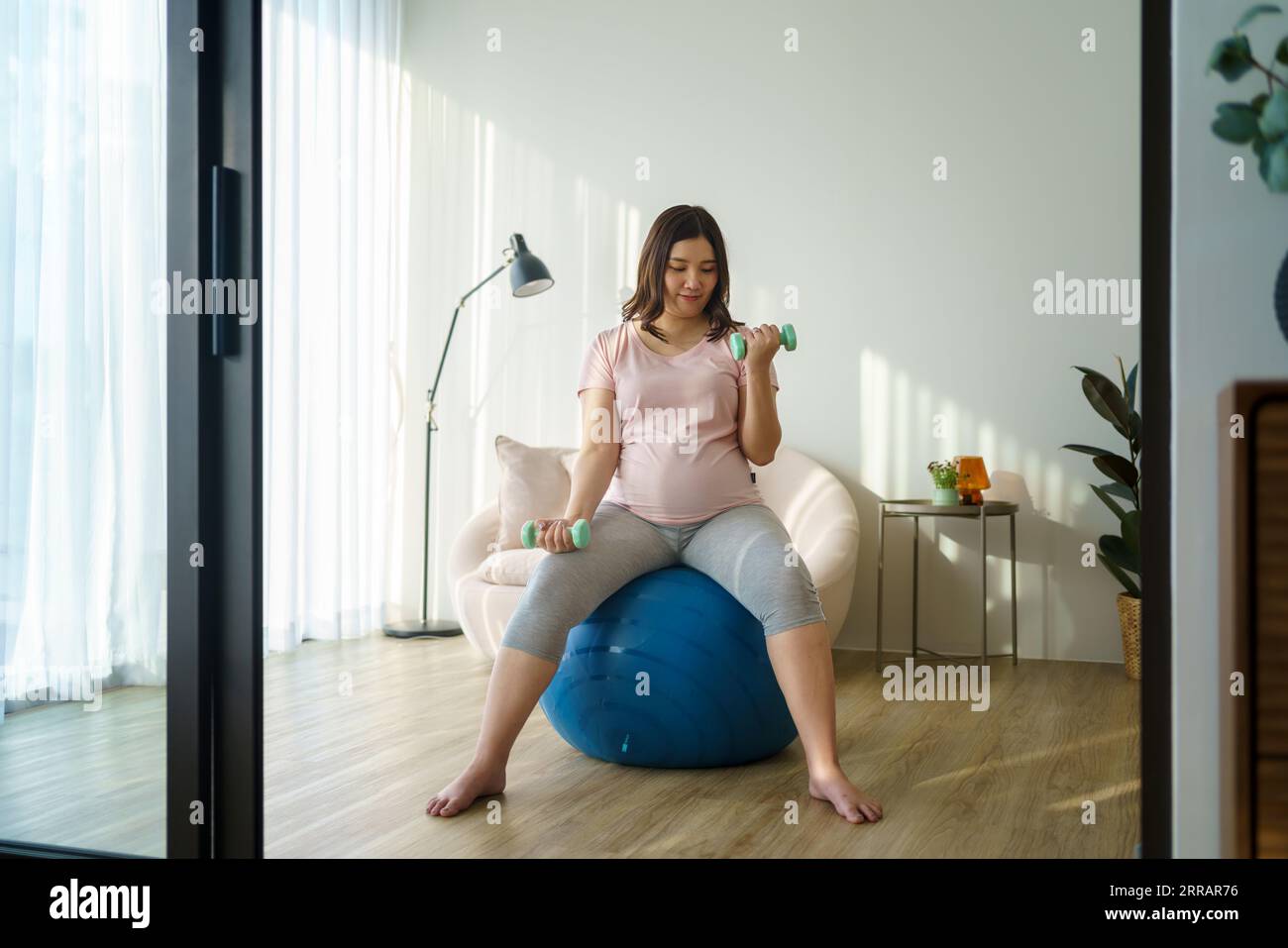 Strahlende asiatische Mom-to-be umarmt Wellness und formt Kraft in ihrem Heim Heiligtum. Kurzhanteln werden zu Werkzeugen der Ermächtigung auf ihrer anmutigen Reise. Stockfoto