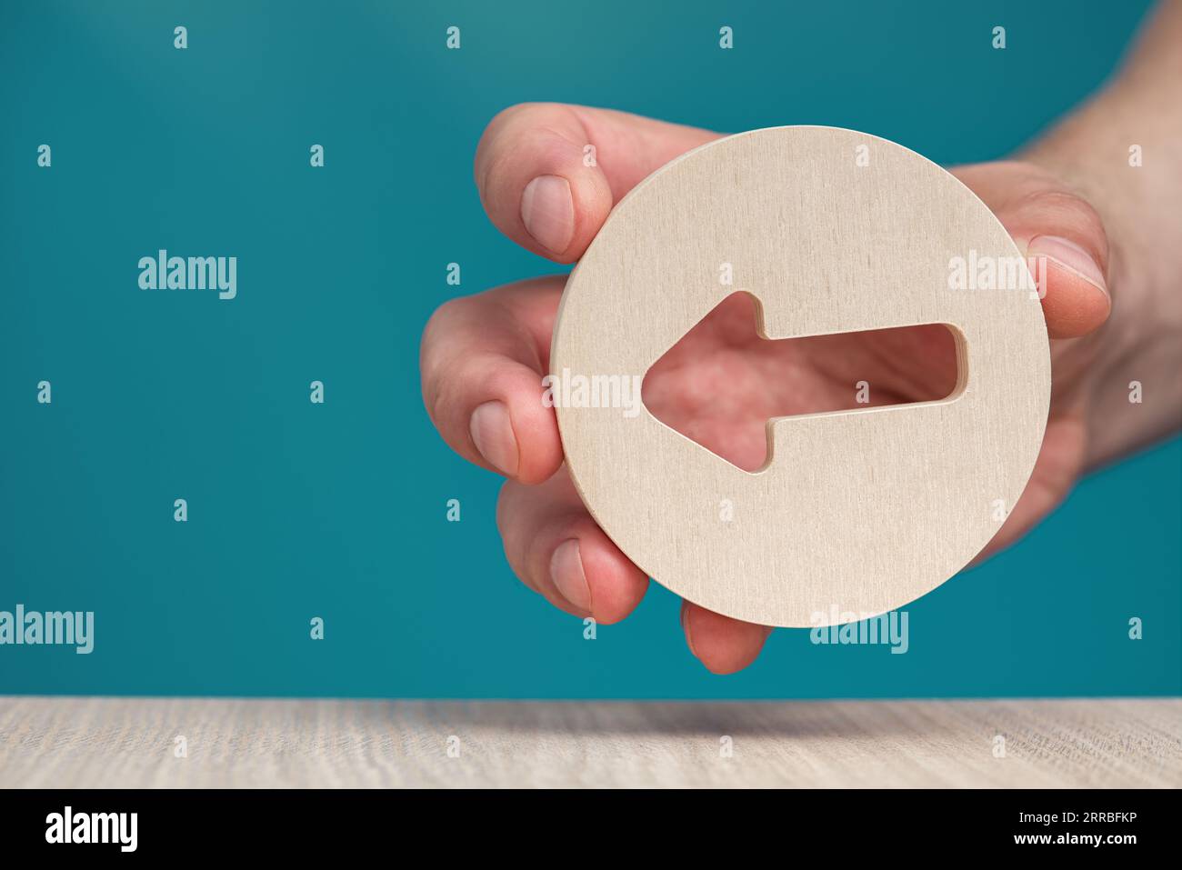 Pfeil zeigt auf blauem Hintergrund nach links. Banner mit einem Pfeil in einem hölzernen Kreis in einer Hand Nahaufnahme zeigt nach links mit Kopierraum. Stockfoto