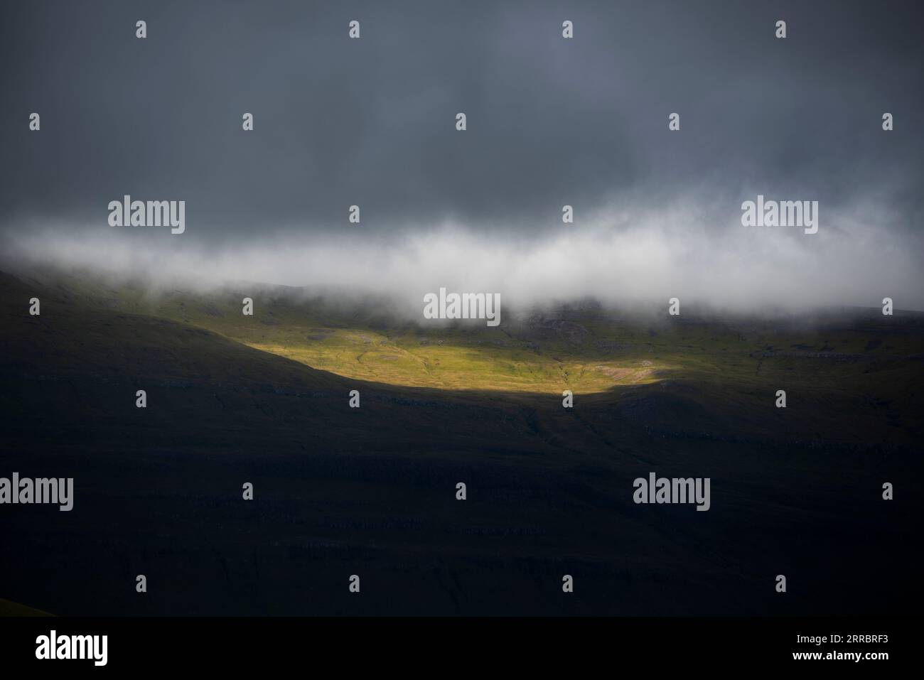 Ein Lichtfleck entweicht der dichten Wolkenbedeckung, um einen abgelegenen Berghang auf den Färöern zu baden. Stockfoto