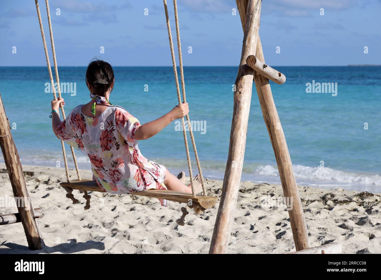 Eine Frau in einem Kleid, die auf einer Holzschaukel gegen das blaue Meer schwingt. Strandurlaub im Resort, Konzept von Freiheit, Glück und Romantik Stockfoto