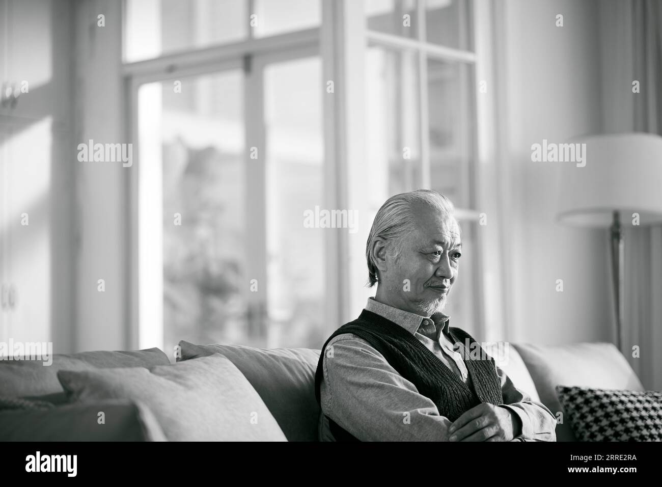 Trauriger asiatischer alter Mann, der allein auf der Couch im Wohnzimmer zu Hause sitzt, schwarz-weiß Stockfoto