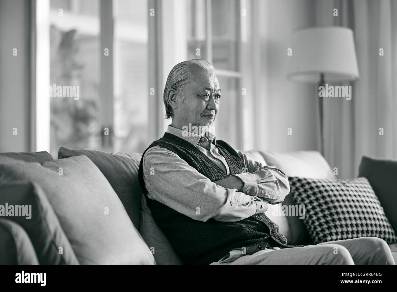 Trauriger asiatischer alter Mann, der allein auf der Couch im Wohnzimmer zu Hause sitzt, schwarz-weiß Stockfoto