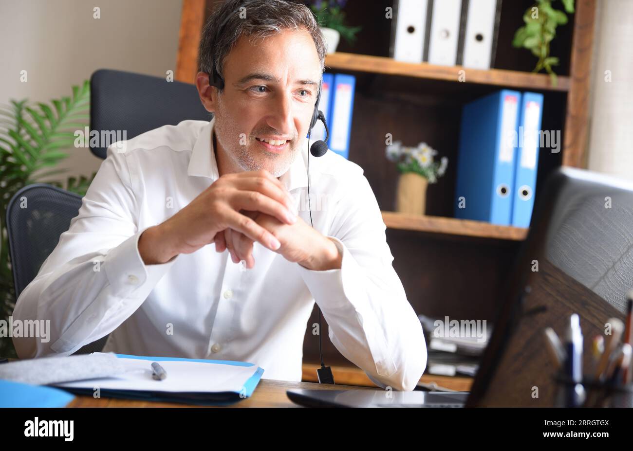 Lächelnder Mann im weißen Hemd Videokonferenzen von einem Laptop aus, der mit Kopfhörern in einem Büro mit einem Tisch und Holzregalen mit Ordnern hört Stockfoto