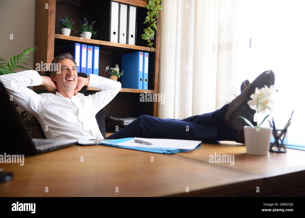 Mann sitzt glücklich in seinem Büro mit seinen Füßen auf dem Holztisch mit Dokumenten und Regalen mit Aktenschränken dahinter Stockfoto
