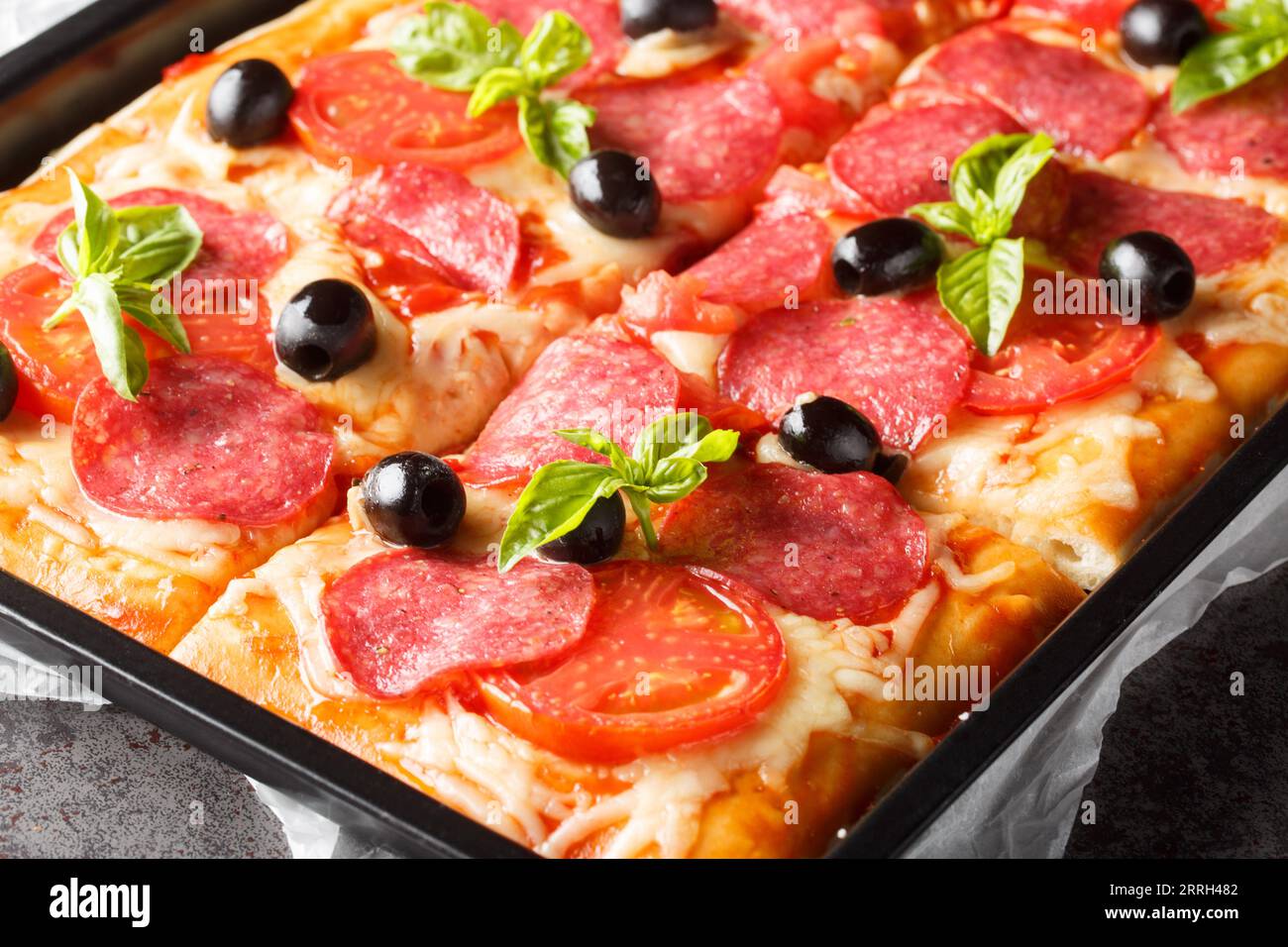 Hausgemachte rechteckige Pizza in Scheiben mit Mozzarella, Salami, Oliven und Tomaten aus nächster Nähe auf einem Backblech auf dem Tisch. Horizontal Stockfoto