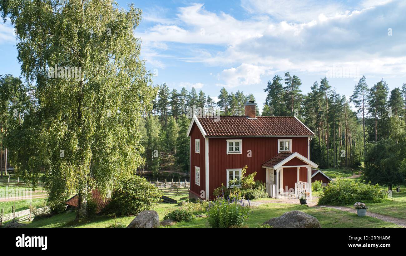 Ein typisches rot-weißes schwedisches Haus im Kleinen. Grüne Wiese im Vordergrund, kleiner Wald im Hintergrund. Blauer Himmel mit kleinen Wolken. Skandinavien lan Stockfoto