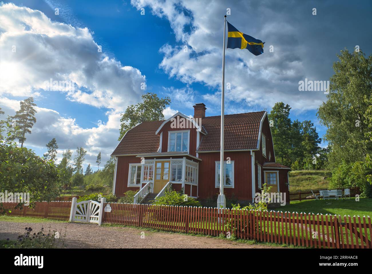 Ein typisches rot-weißes schwedisches Haus im Kleinen. Weißes Gartentor, brauner Zaun. Fahnenmast mit schwedischer Flagge. Grüner Rasen im Garten. Bäume im Hintergrund Stockfoto
