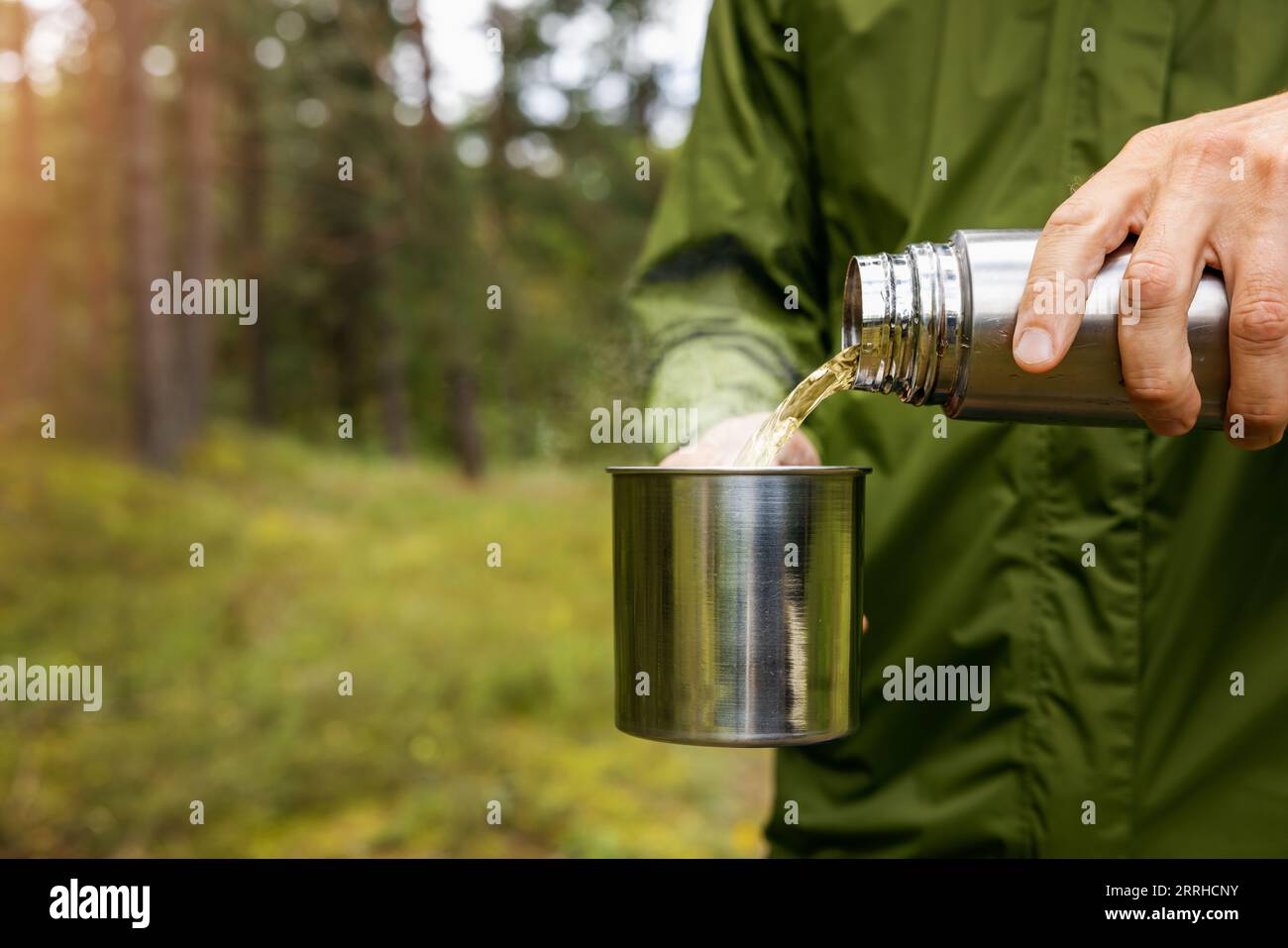 Der Mensch gießt ein heißes Getränk aus Thermoskanne in einen Metallbecher im Wald. Naturtourismus und Campingausrüstung. Leerzeichen kopieren Stockfoto