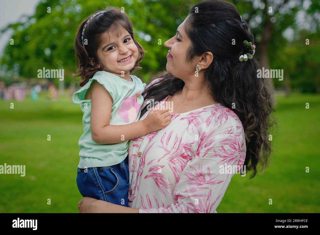 Glückliche verspielte junge indische Mutter und süße kleine Mädchentochter im Sommerpark oder Garten. Stockfoto