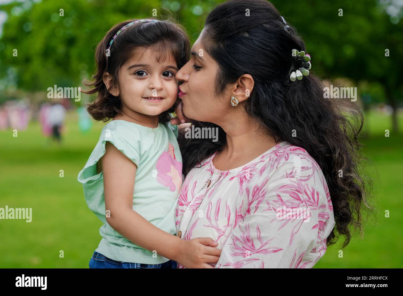 Glückliche junge indische Mutter küsst ihre süße kleine Tochter im Sommerpark. Stockfoto