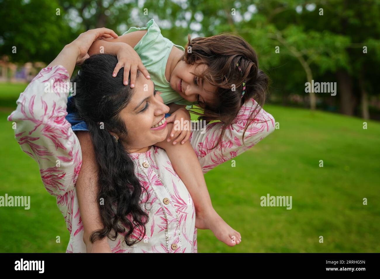 Glückliche junge indische Mutter, die ihre Tochter im Sommerpark auf den Schultern trägt. Familien- und Mutterschaftskonzept. Stockfoto