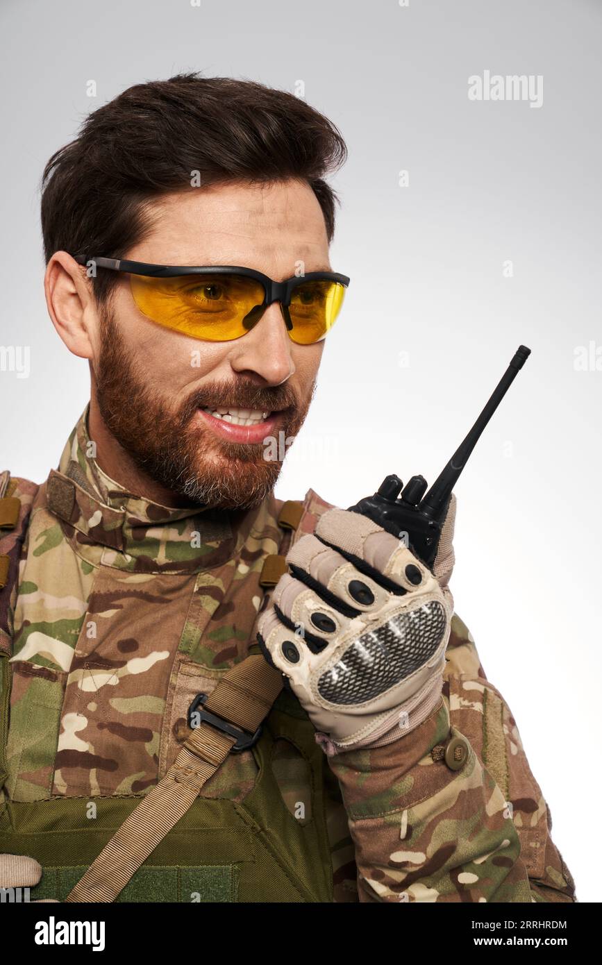 Gut aussehender Militärmann in gelben taktischen Gläsern, der mit Walkie-Talkie spricht. Porträt eines lächelnden Soldaten in Tarnuniform, mit tragbarem Radio, Berichterstattung, isoliert auf weiß. Verbindungskonzept. Stockfoto